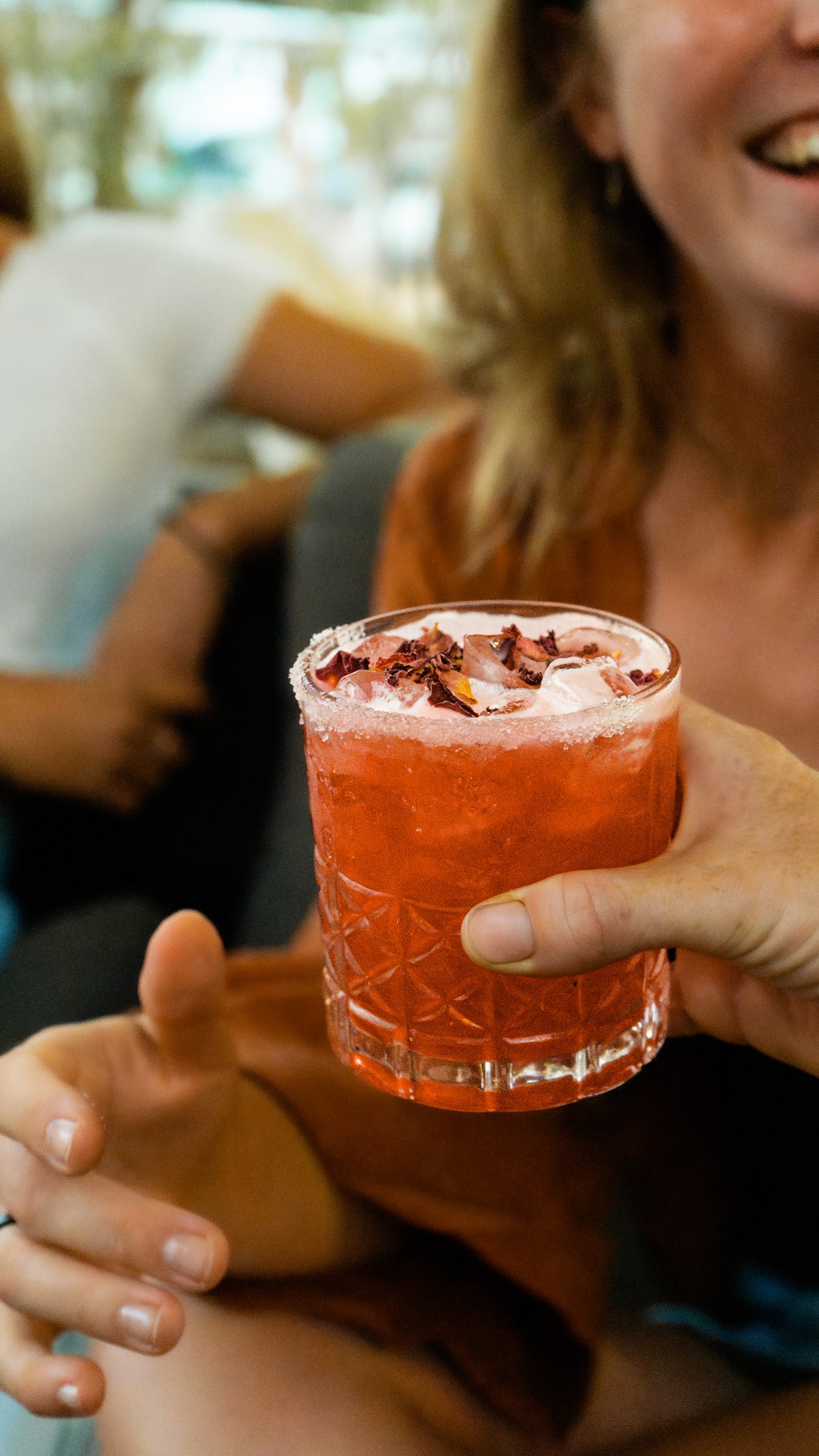 Hibiscus Cocktail at Mutts Café Close-up of hibiscus cocktail with a girl reaching for it, sunset and sunny summer vibes, blurred background. Drinks Menu Exmouth