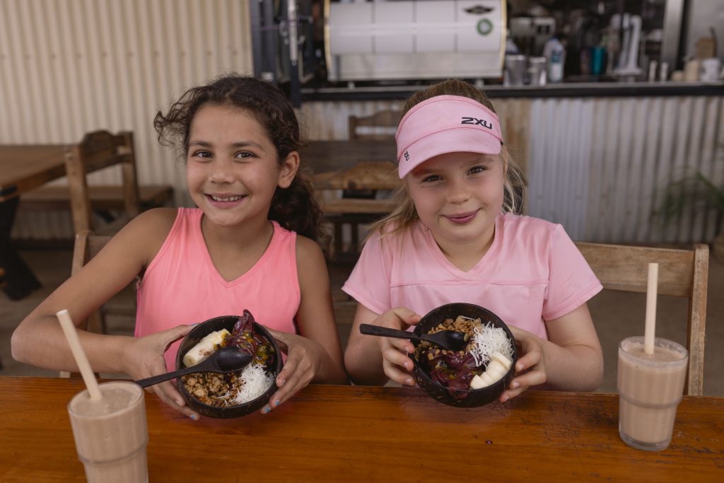Açaí Bowls for Kids & Families | Mutts Café Exmouth Two smiling kids holding açaí bowls outside at Mutts Café Exmouth with smoothies, enjoying breakfast in the sunshine.