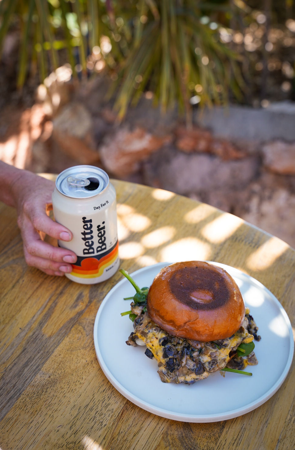 Better Beer with Mushroom Burger at Mutts Café Person holding a Better Beer next to a mushroom burger, outside in the garden at Mutts Café on a sunny day. Drinks Menu Exmouth