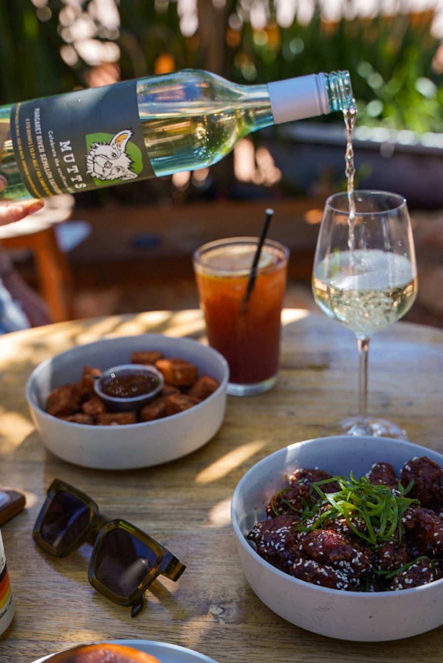 Mutts Café White Wine Pour White wine being poured into a glass with Mutts label, food on table in the background, outside in the garden. Drinks Menu Exmouth