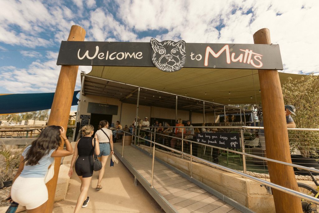 People walking into Mutts Café Exmouth under a large “Welcome to Mutts” sign on a sunny day in Exmouth WA. Contact Mutts Café Exmouth