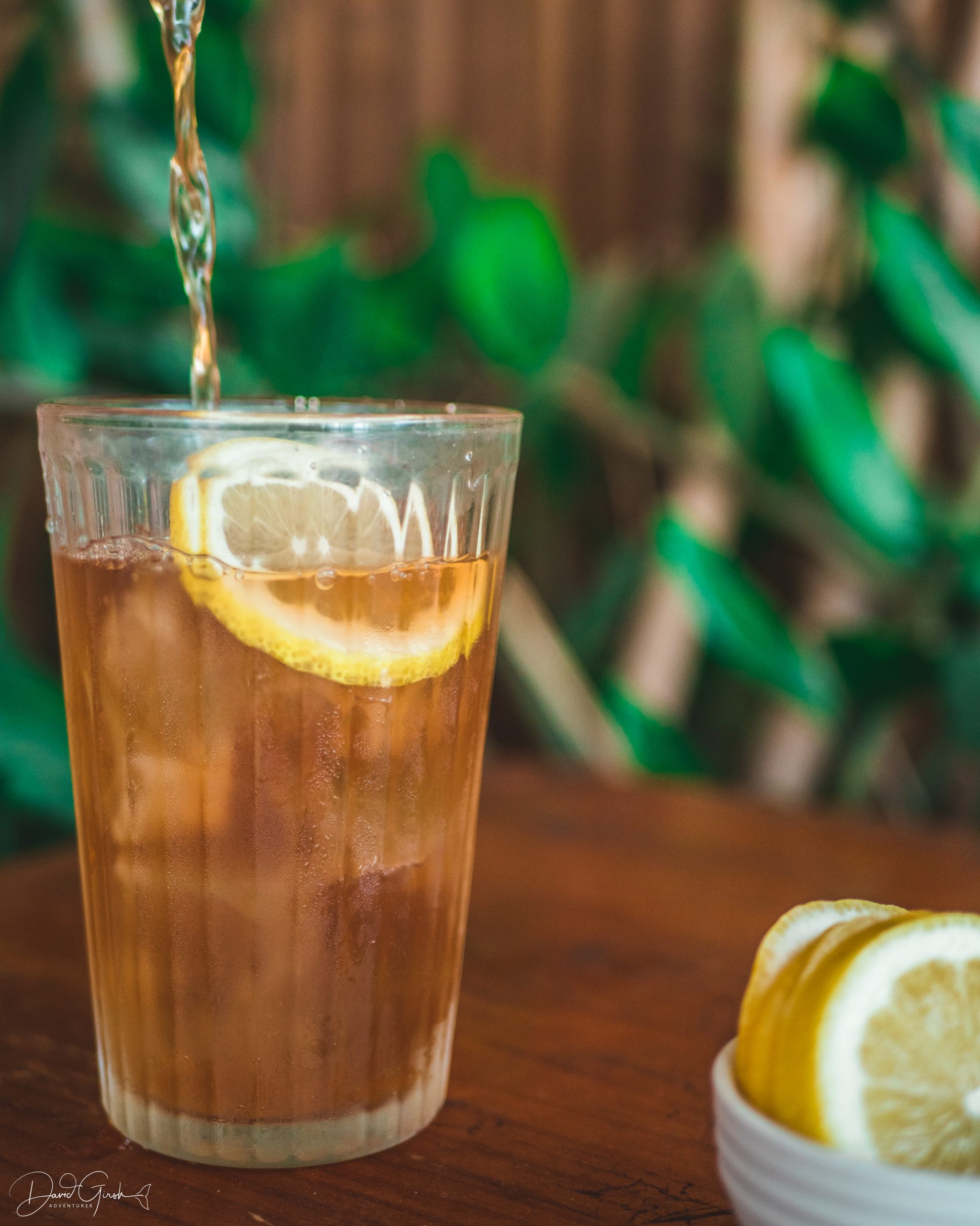 Action shot of lemon iced tea being poured into a glass, lemons in foreground, outdoors at Mutts Café with greenery in the background. Drinks Menu Exmouth